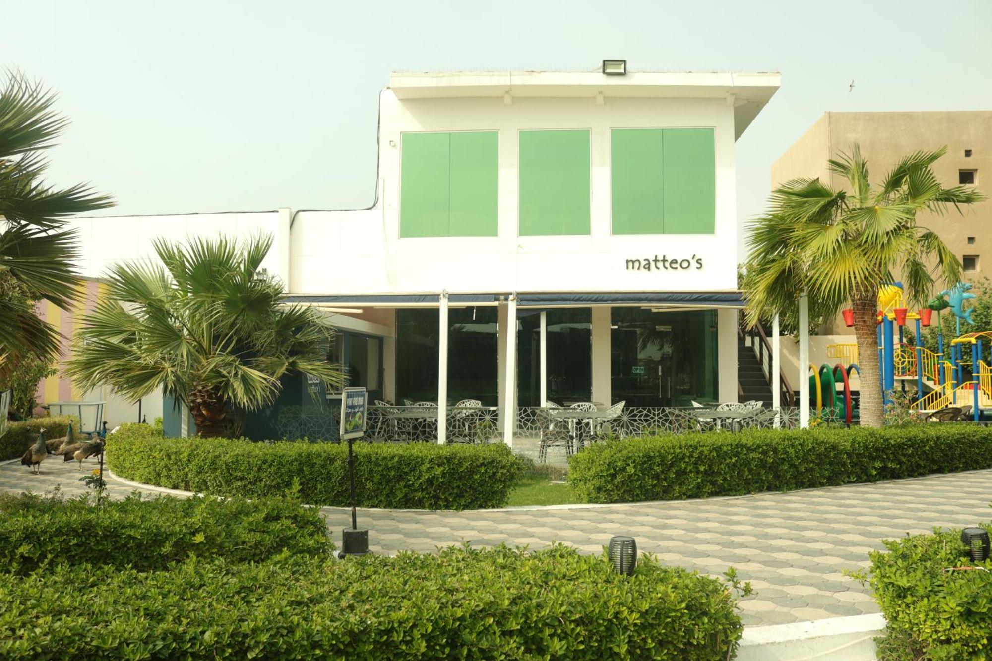 Lagoona Beach Luxury Resort And Spa Manama Exterior foto The photo shows a building with the name "matteo's" displayed prominently on the front. The structure features large windows and green shutters. There is a landscaped area in front with bushes and palm trees, along with a paved pathway leading up to 