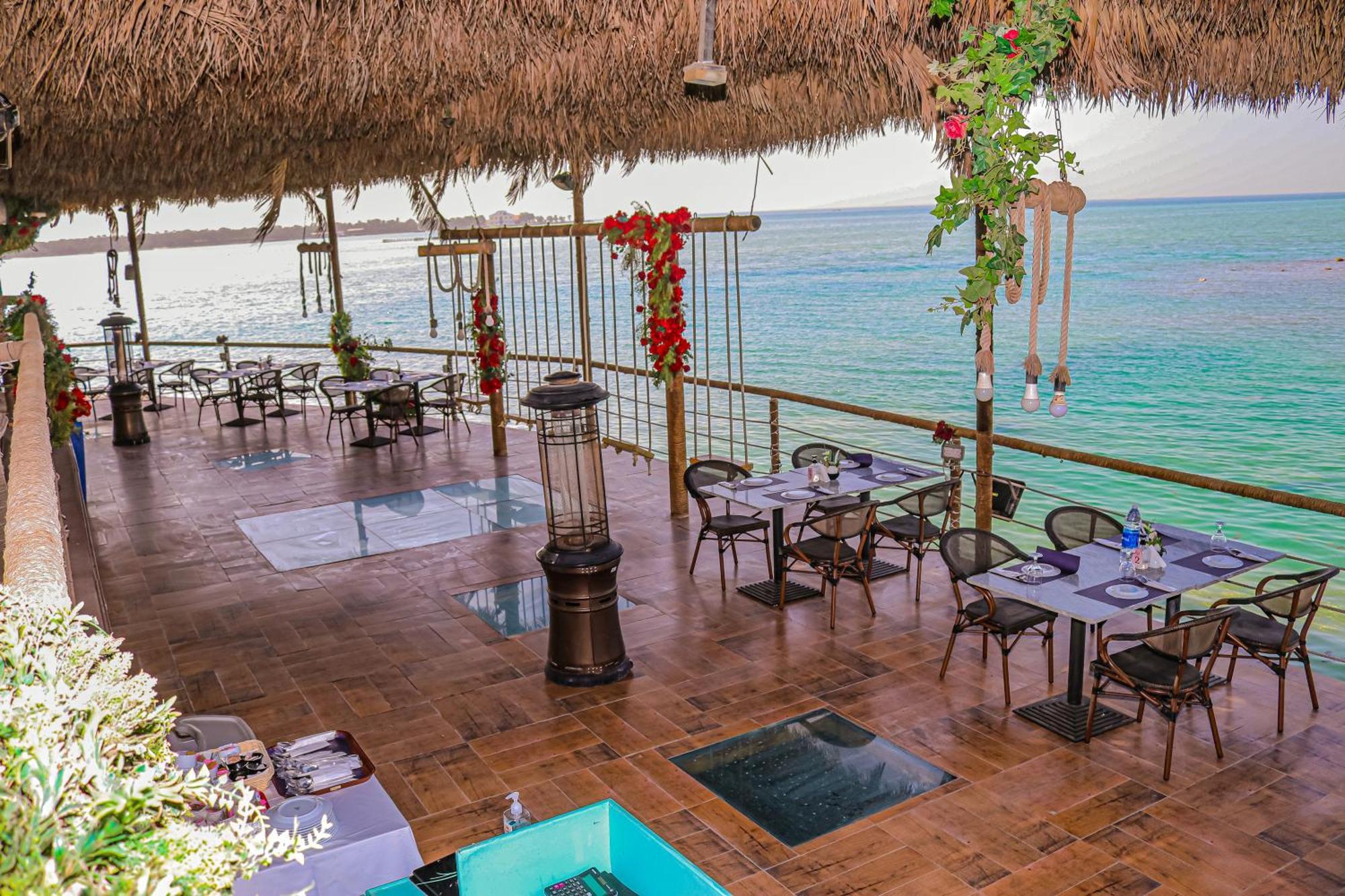 Lagoona Beach Luxury Resort And Spa Manama Exterior foto The photo depicts a coastal restaurant or dining area with an open-air design. It features a wooden floor and tables set for dining, surrounded by chairs. The space is partially covered by a thatched roof, providing shade while allowing for a view of