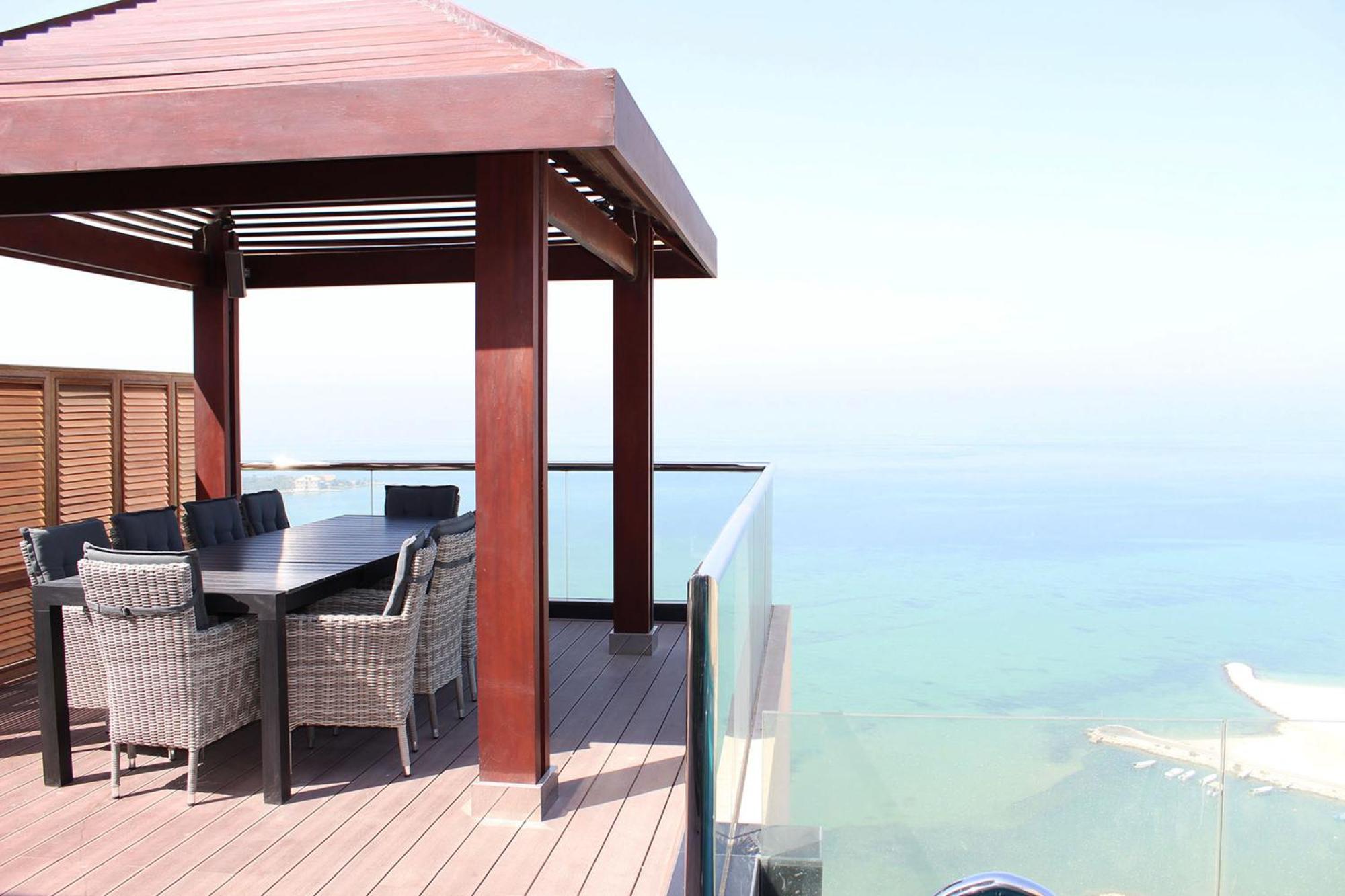 Lagoona Beach Luxury Resort And Spa Manama Exterior foto The photo showcases a stylish outdoor patio or terrace area. It features a wooden structure with a sloped roof, providing a shaded space. There is a large table made of dark wood, surrounded by several wicker chairs. The patio is elevated and has gla