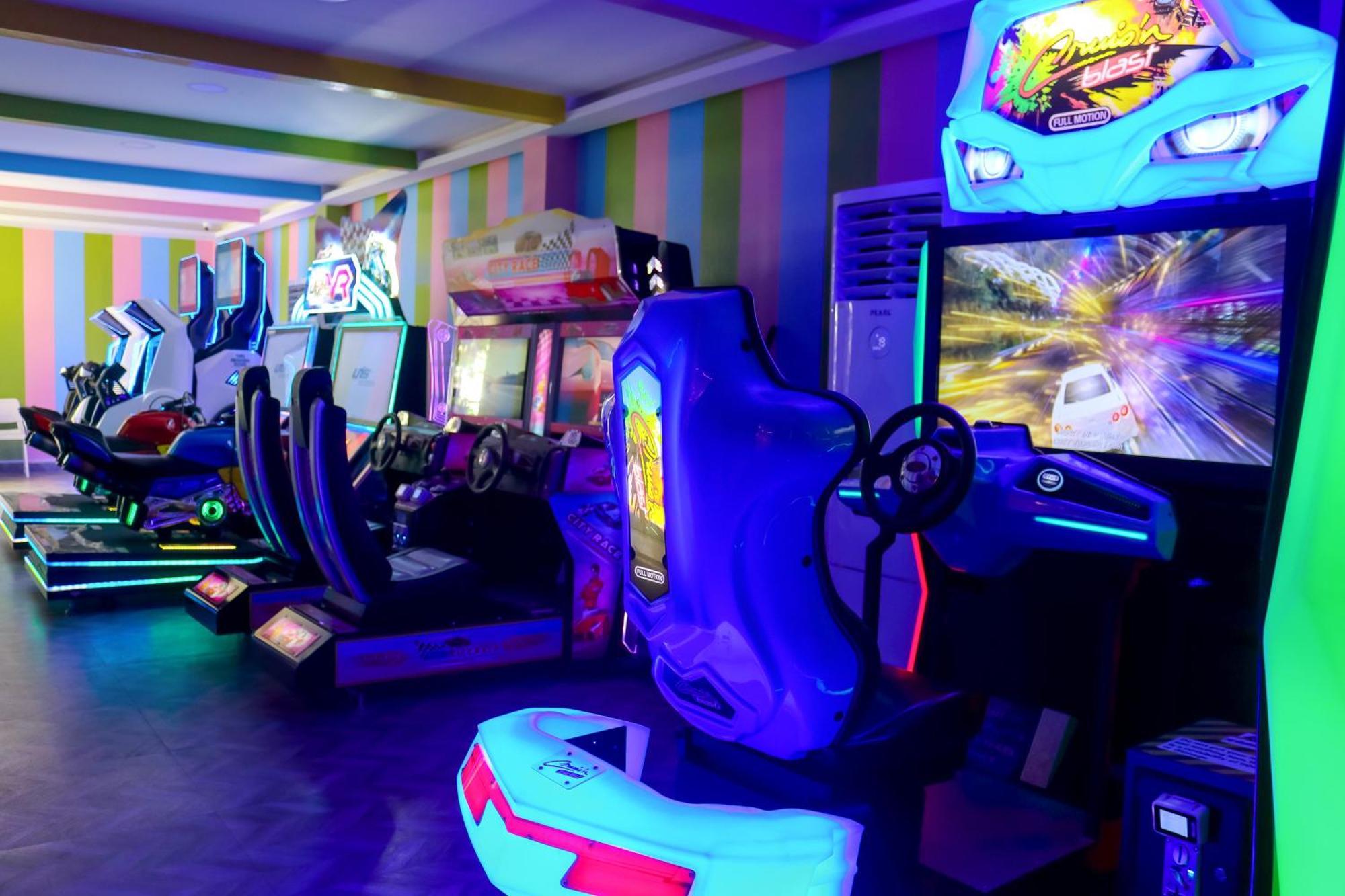 Lagoona Beach Luxury Resort And Spa Manama Exterior foto The photo shows an arcade area filled with various arcade machines. The setting is illuminated with colorful neon lights, creating a vibrant atmosphere. There are several racing game machines with steering wheels and seats, each accompanied by screen