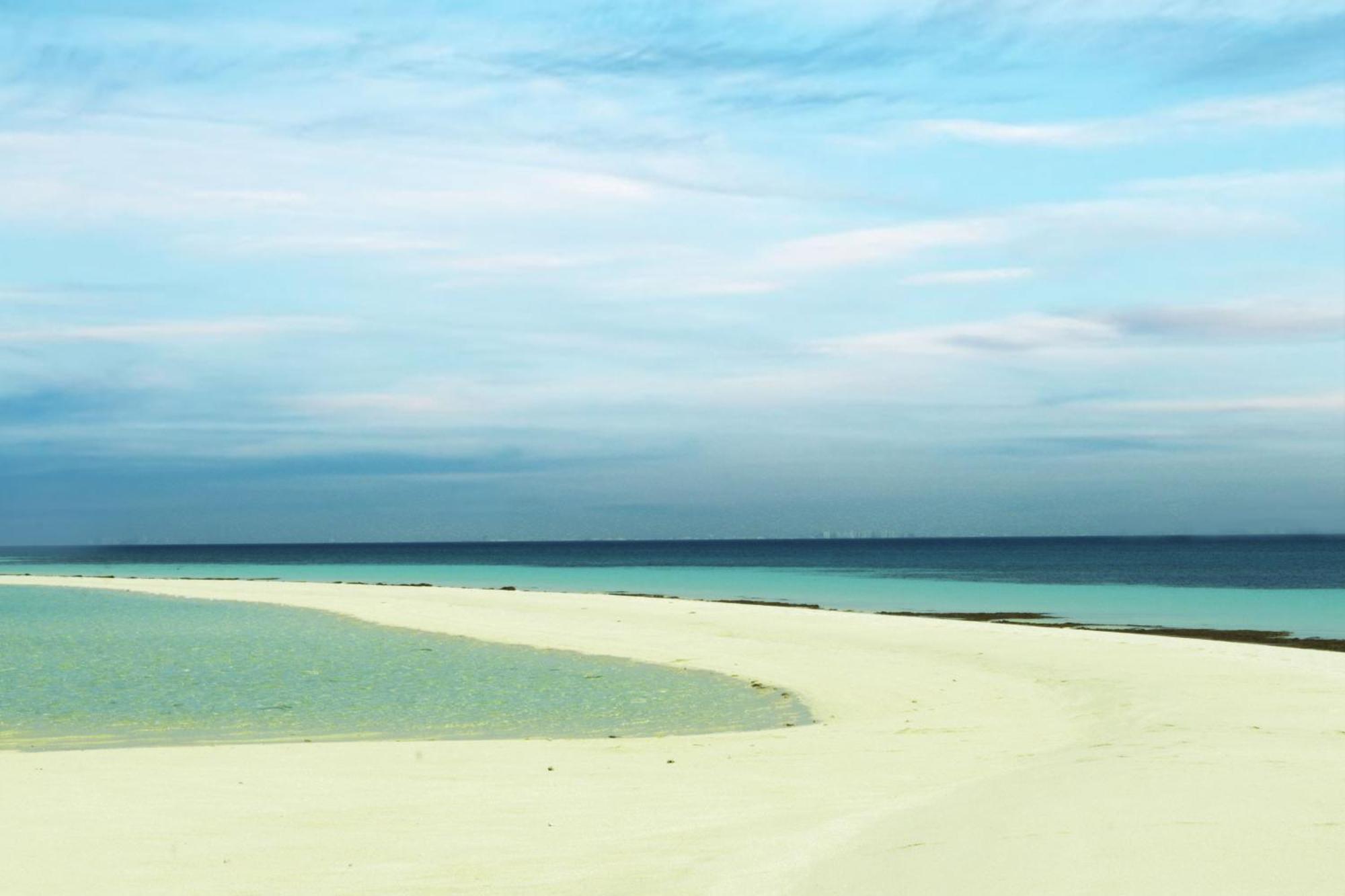 Lagoona Beach Luxury Resort And Spa Manama Exterior foto The photo depicts a serene beach scene with a gentle curve of sandy shore leading into shallow turquoise waters. The calm ocean stretches into the horizon, meeting a sky filled with soft clouds in various shades of blue. The overall atmosphere is tra