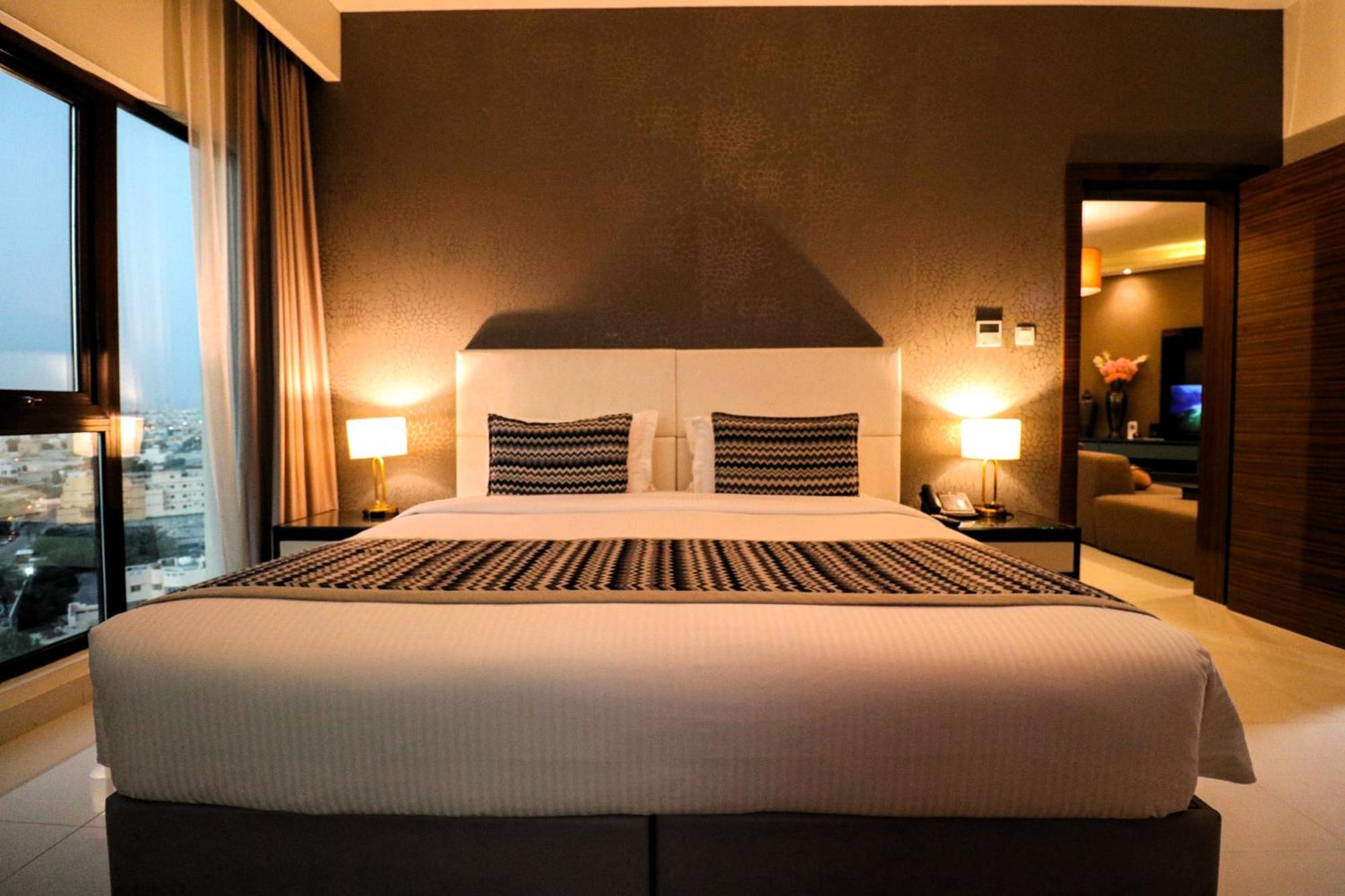 Lagoona Beach Luxury Resort And Spa Manama Exterior foto The photo depicts a modern hotel room featuring a large bed with a white comforter and two decorative pillows. The headboard is light-colored, and the walls are painted a dark hue. On either side of the bed, there are bedside tables with lamps. The r