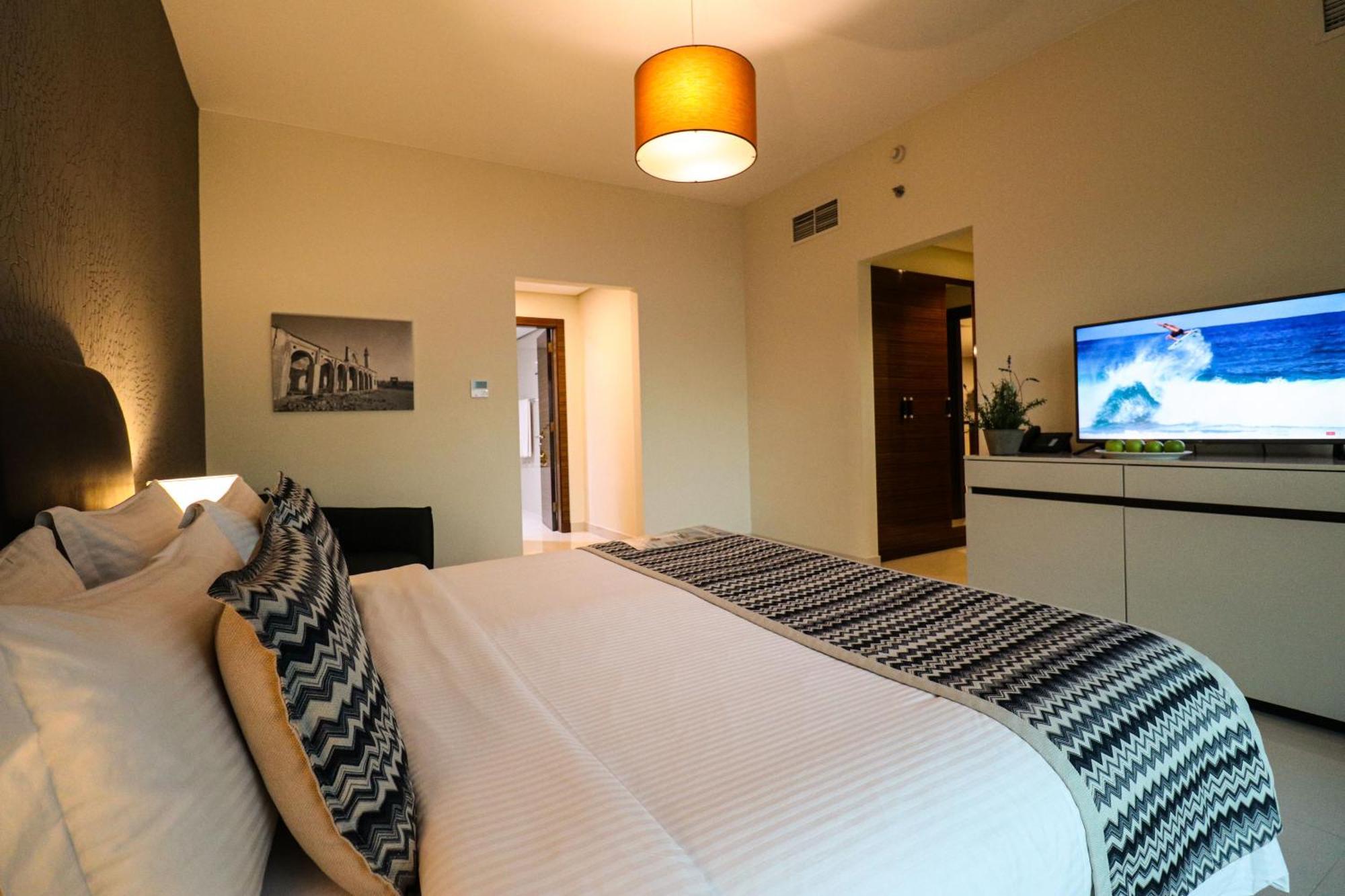 Lagoona Beach Luxury Resort And Spa Manama Exterior foto The photo shows a well-decorated hotel room. There is a large bed with white linens and decorative pillows featuring a zigzag pattern. On the wall beside the bed is a piece of artwork, and there is a modern television on a white furniture unit opposi