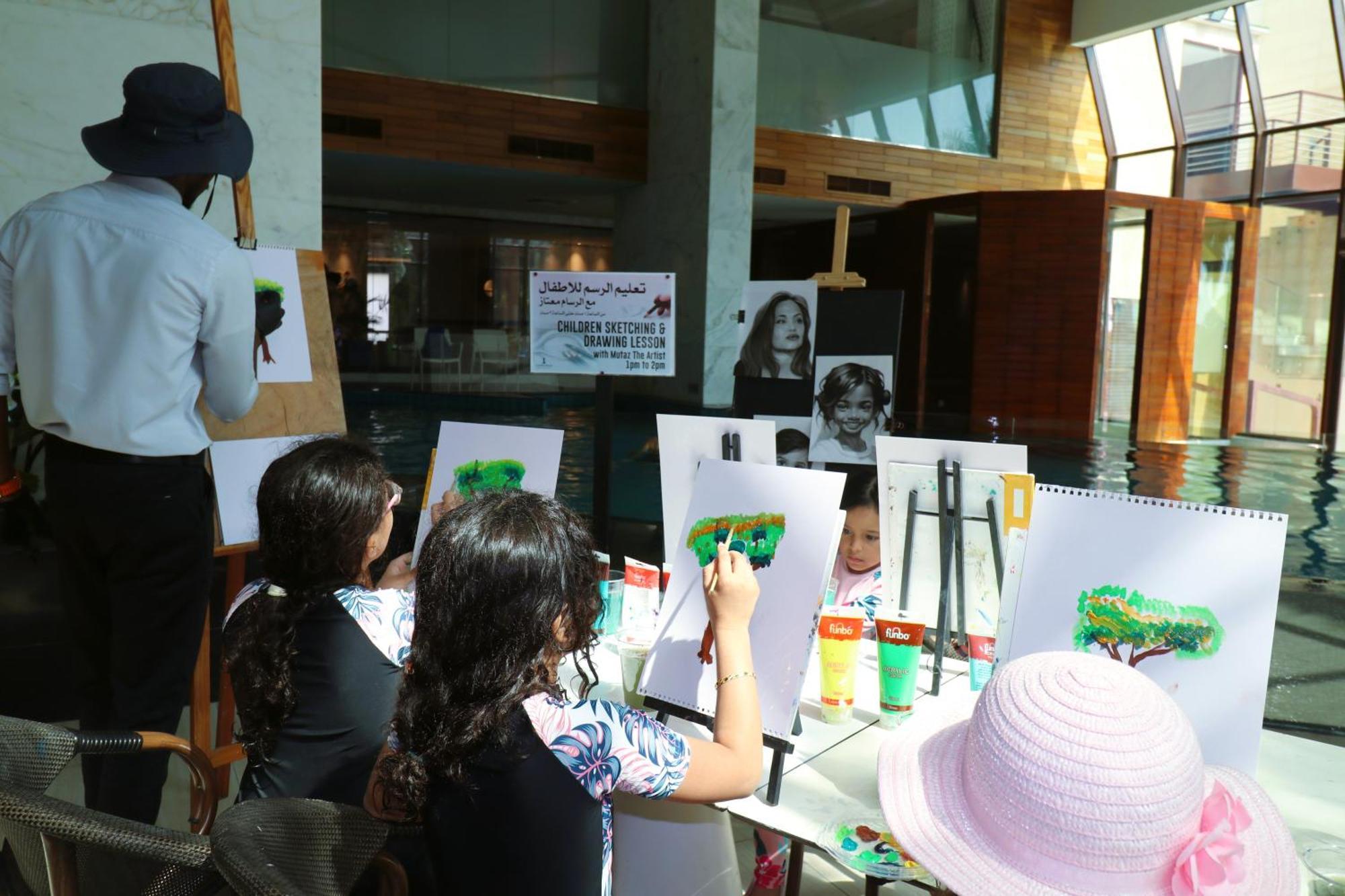 Lagoona Beach Luxury Resort And Spa Manama Exterior foto The photo shows a group of children and an adult engaged in an art activity near a pool area. The children are seated at a table, painting on canvases or paper, with a focus on trees, as indicated by the green paint they are using. An adult, wearing 